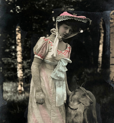 Zdjęcie koloryzowane z początku XX wieku. Młoda kobieta w letniej eleganckiej sukni z kapeluszem na głowie, przewiązanym pod brodą. Na dłoniach ma koronkowe rękawiczki. Obok stoi dog