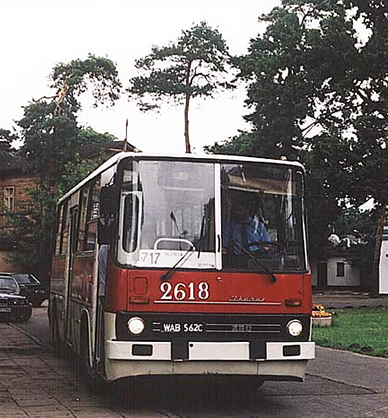 Fotografia kolorowa. Pętla autobusowa. W centralnym miejscu stoi miejski autobus pomalowany na czerwony i biały kolor. Po lewej stronie na chodniku kilka osób z rowerami, po prawej niski budynek, trawa i drzewa