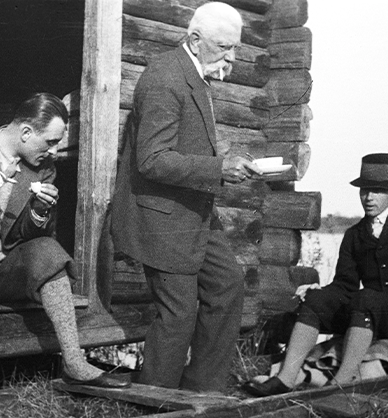 Fotografia czarno-biała. Trzech mężczyzn odpoczywających w kureniu, drewnianym budynku mieszkalnym, na Polesiu. Ubrani w męskie garnitury. Dwóch z nich ma założone skarpety do wysokości łydek. Jeden siedzi u wejścia do chaty, dwóch jest na łódce. Mężczyzna w środku trzyma filiżankę w dłoni