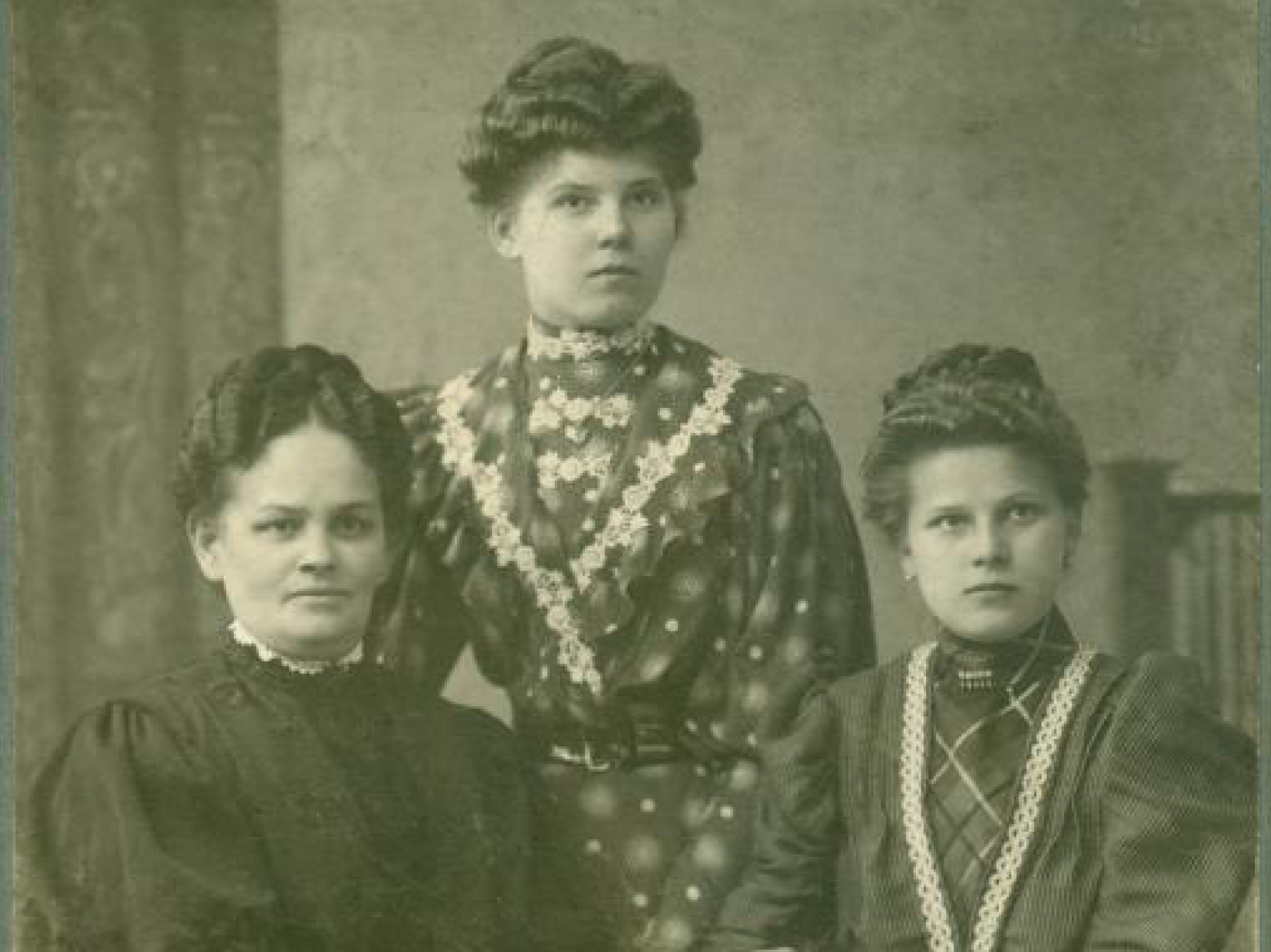 Portret trzech kobiet z początku XX wieku, ubranych w eleganckie długie suknie. Wszystkie kobiety mają upięte włosy. Dwie siedzą przy stole, jedna stoi pomiędzy nimi. Na stole leży obrus i stare pisma. Zdjęcie wykonane w atelier fotograficznym.  Link do strony z artykułem zainspirowanym Zbiorami Społecznymi