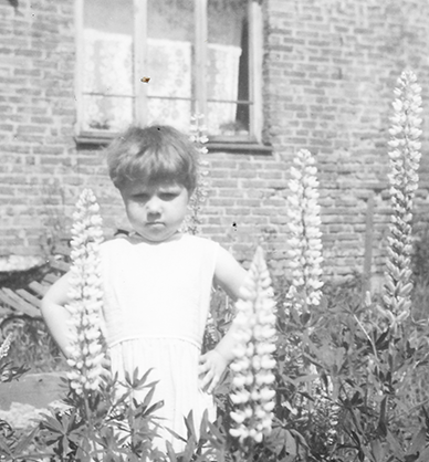 Fotografia czarno-biała. Kilkuletnia dziewczynka stoi w ogrodzie przed domem. Ma nadąsany wyraz twarzy i podpiera się pod boki. Dom z murowanej cegły z dużymi oknami. W jednym oknie wisi biała firanka. W ogrodzie kwitnie bujna trawa i rosną kwiaty łubinu
