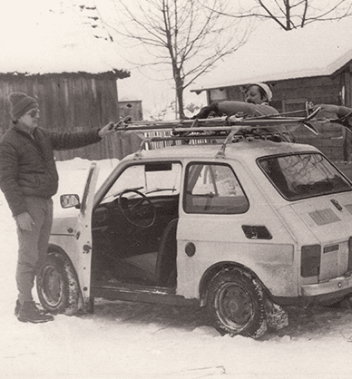 Zima. Dookoła leży śnieg. Trzech mężczyzn stojących przy samochodzie Fiat 126p. Przypinają narty do bagażnika dachowego. W tle widać drewniane zabudowania gospodarcze. Link do podstrony z tym zdjęciem