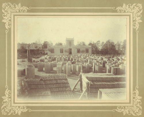 Sepiowa fotografia plenerowego składu ceramiki z rzędami dużych ceramicznych dzbanów i garnków, budynkiem w tle i drzewami w oddali. Ozdobna obwódka otacza obraz.