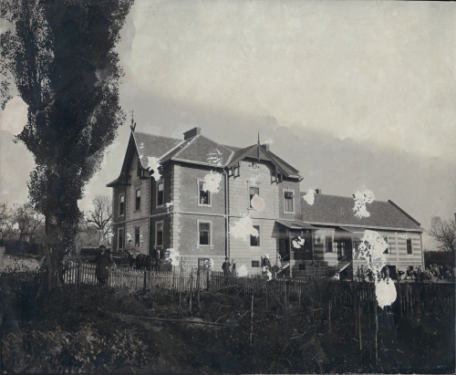 Duży, dwupiętrowy drewniany dom z gankiem i ogrodzonym podwórkiem, otoczony drzewami. Na starej fotografii widoczne są uszkodzenia i plamy.