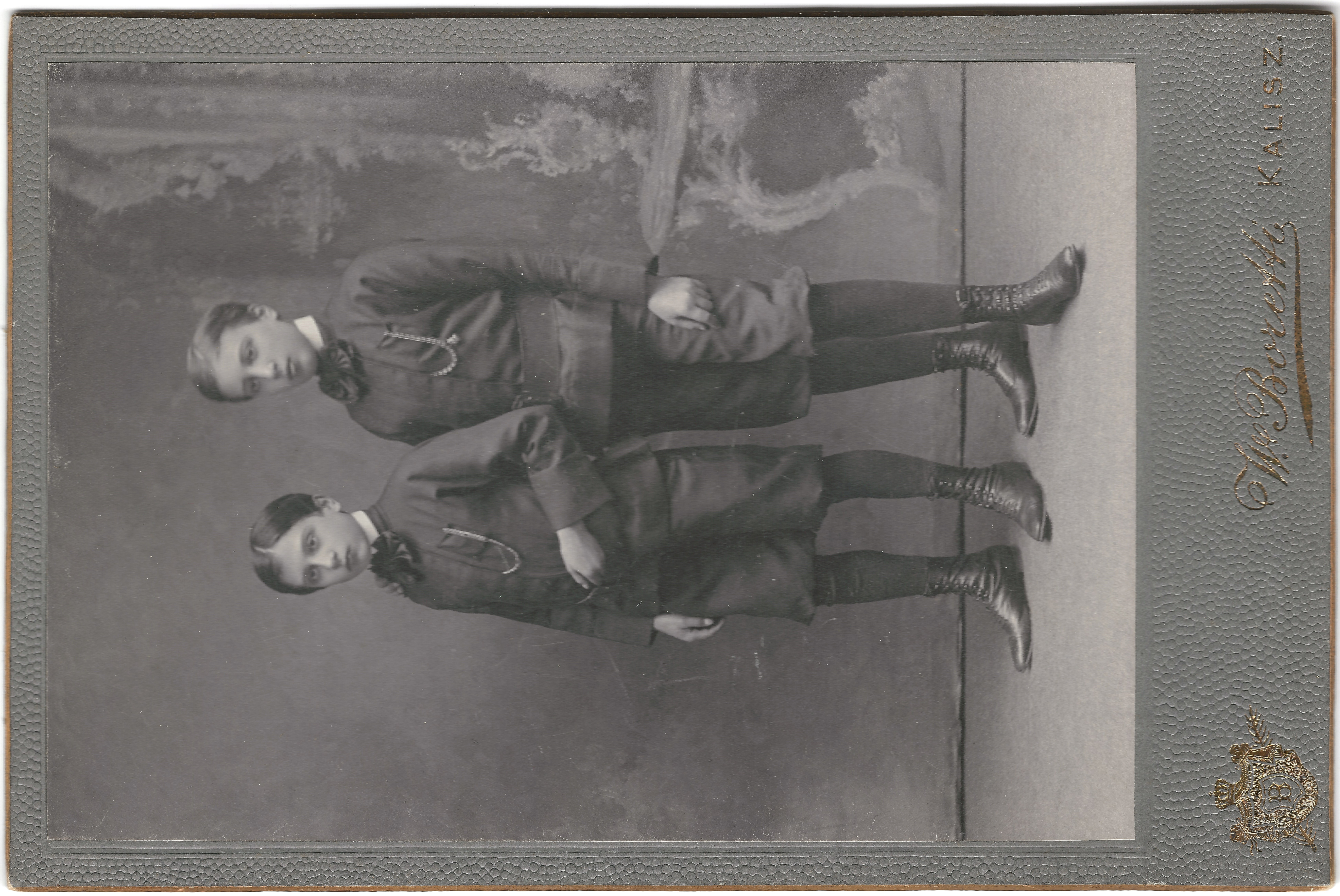 Dwóch chłopców w formalnych strojach z epoki wiktoriańskiej stoi obok siebie na portrecie studyjnym z namalowanym tłem, wytłoczoną obwódką i nazwiskiem fotografa u dołu.