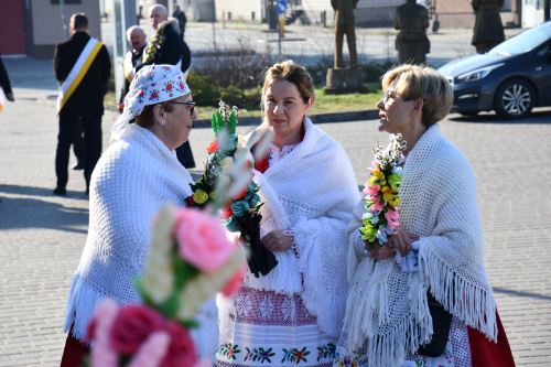 Trzy kobiety w tradycyjnych białych szalach i kwiecistych spódnicach stoją na zewnątrz, trzymają dekoracyjne kompozycje kwiatowe i rozmawiają podczas dziennego wydarzenia.