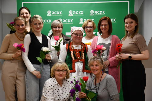 Grupa dziesięciu kobiet, niektóre z nich trzymają kwiaty, pozuje na zielonym tle z logo „RCKX”; jedna kobieta ma na sobie tradycyjny strój ludowy.