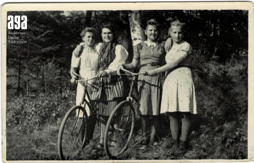 Cztery kobiety w ubraniach vintage stoją razem na zewnątrz, dwie z nich trzymają kierownicę roweru.