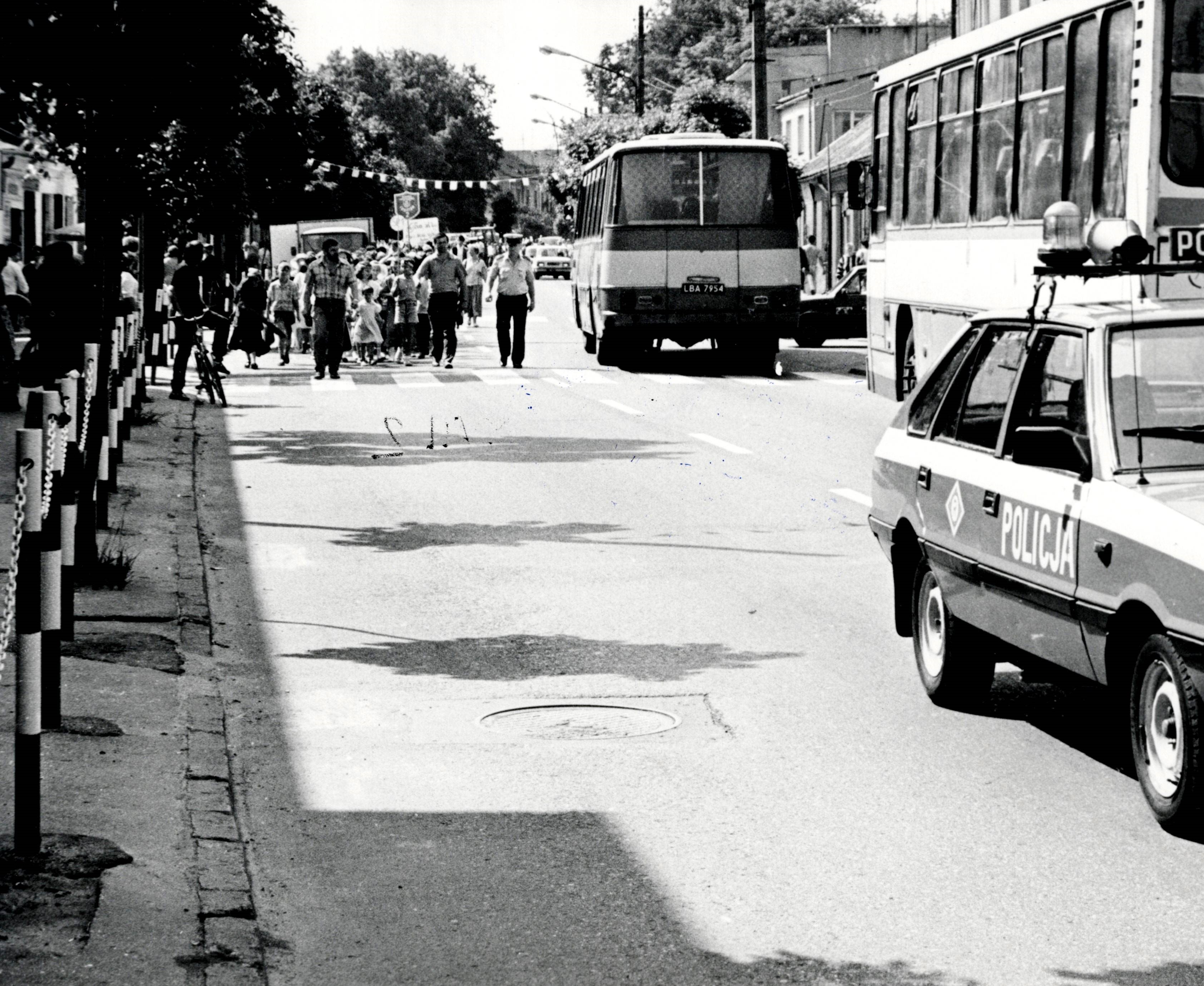 Czarno-białe zdjęcie sceny ulicznej z radiowozem i autobusem. Ludzie chodzą po chodniku, a ruch jest niewielki. Drzewa i budynki rosną wzdłuż drogi.