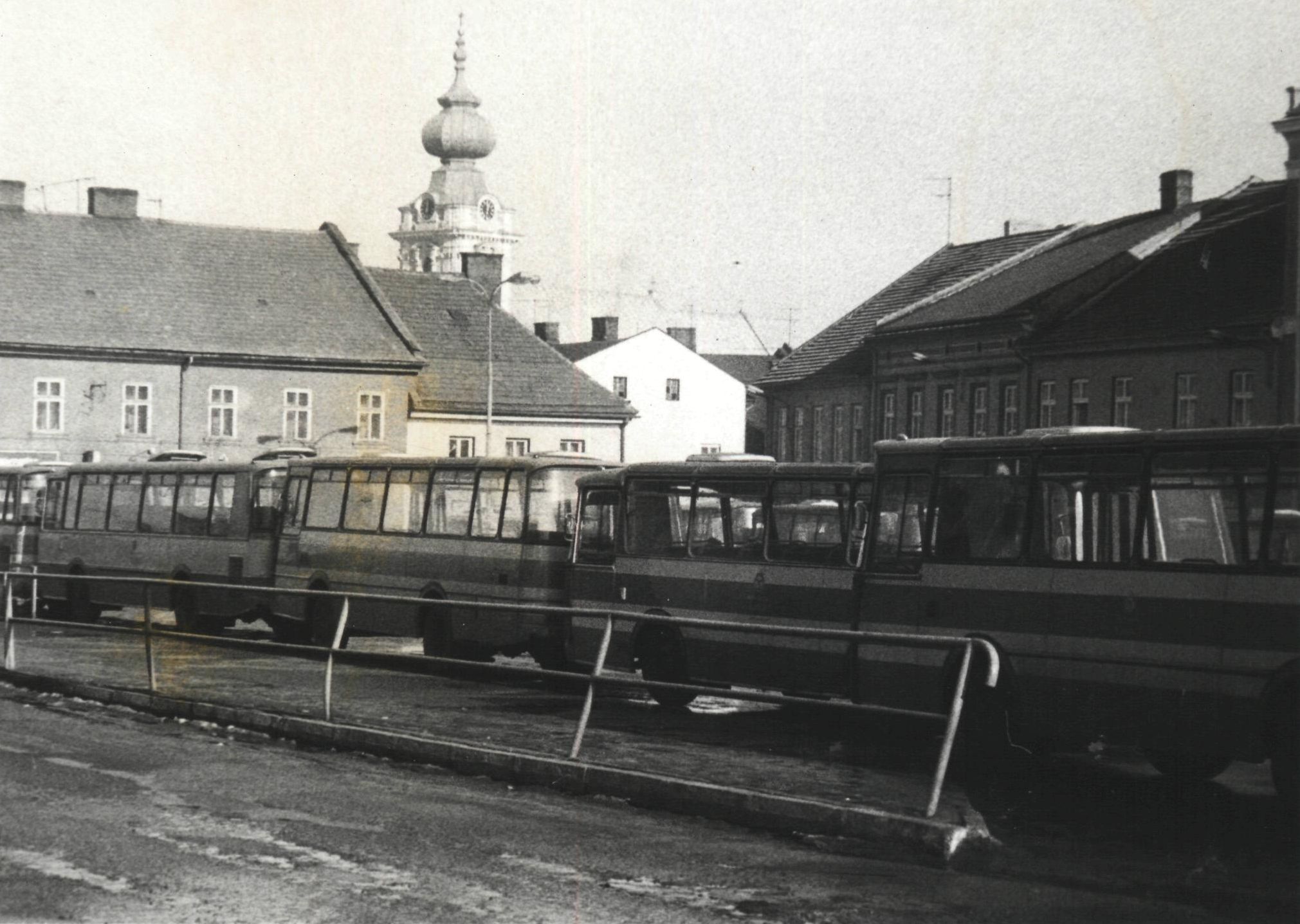 Rząd zaparkowanych autobusów na ulicy. W tle widać stare budynki i kopułę kościoła. Na czarno-białej fotografii.