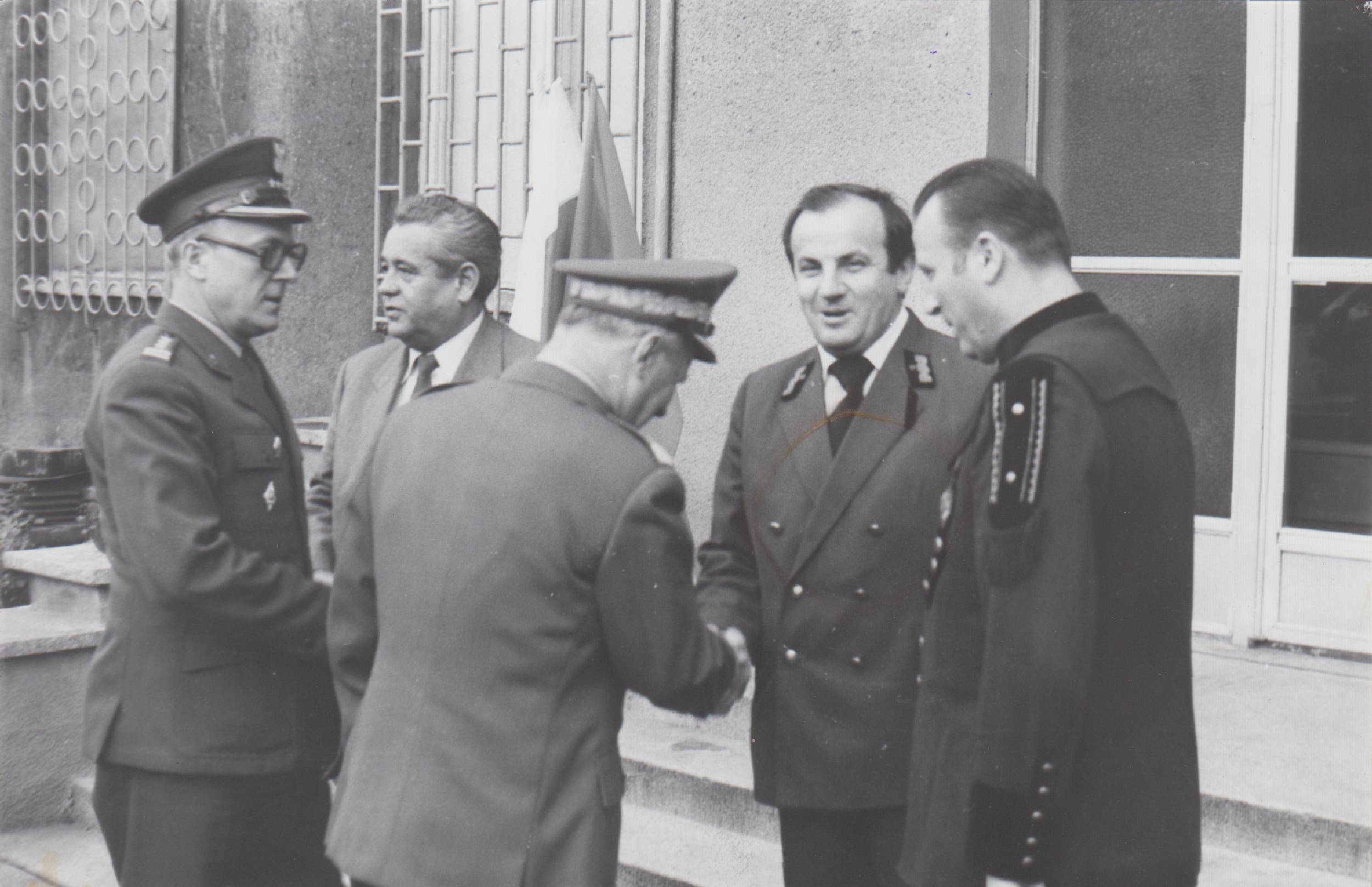 Pięciu mężczyzn ubranych w formalne i wojskowe stroje rozmawia na zewnątrz, dwóch ściska im dłonie.