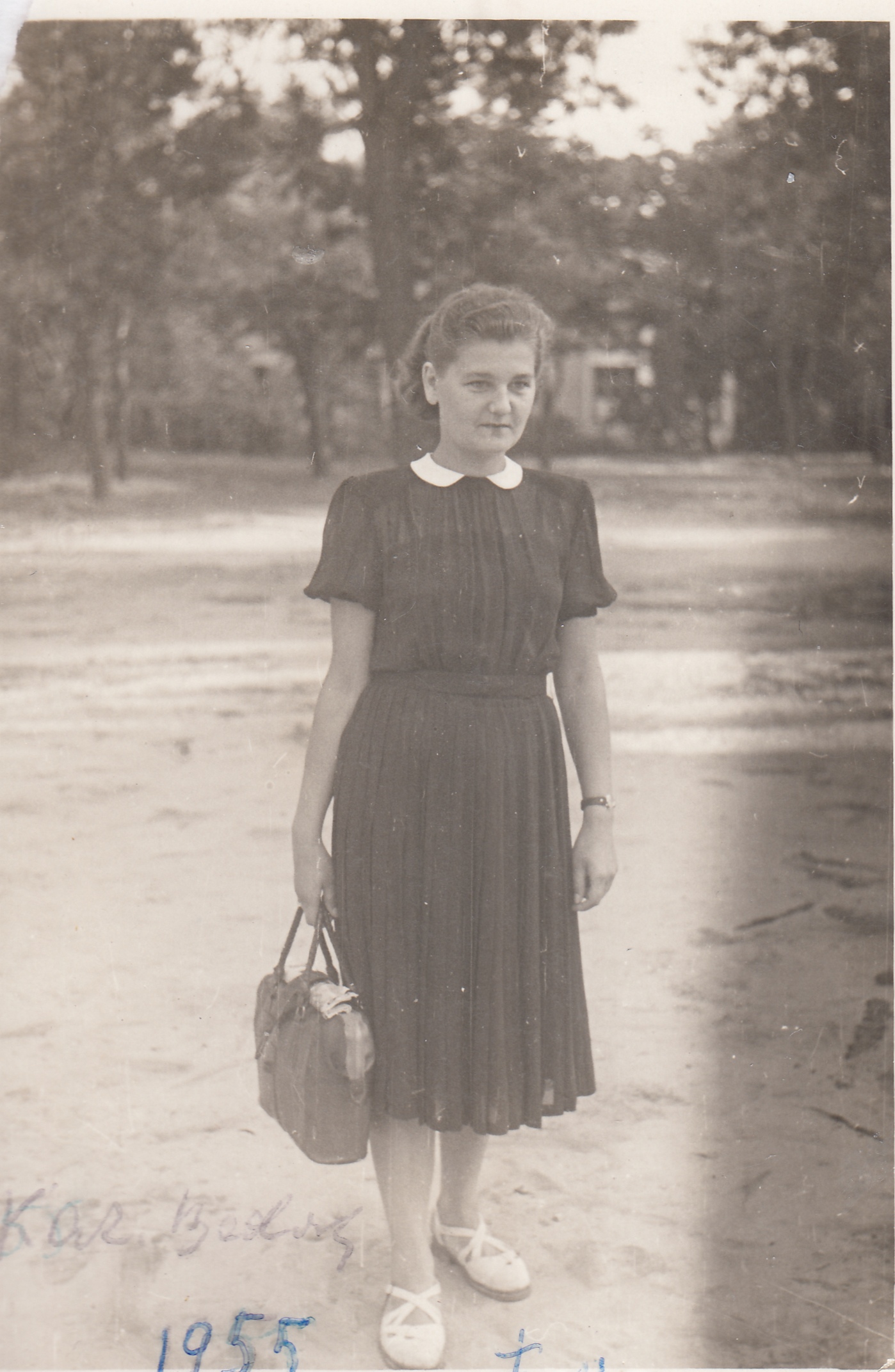 Kobieta w ciemnej sukience stoi na zewnątrz trzymając torebkę. Obraz jest czarno-biały i datowany na 1955 r. W tle widoczne są drzewa i konstrukcja.