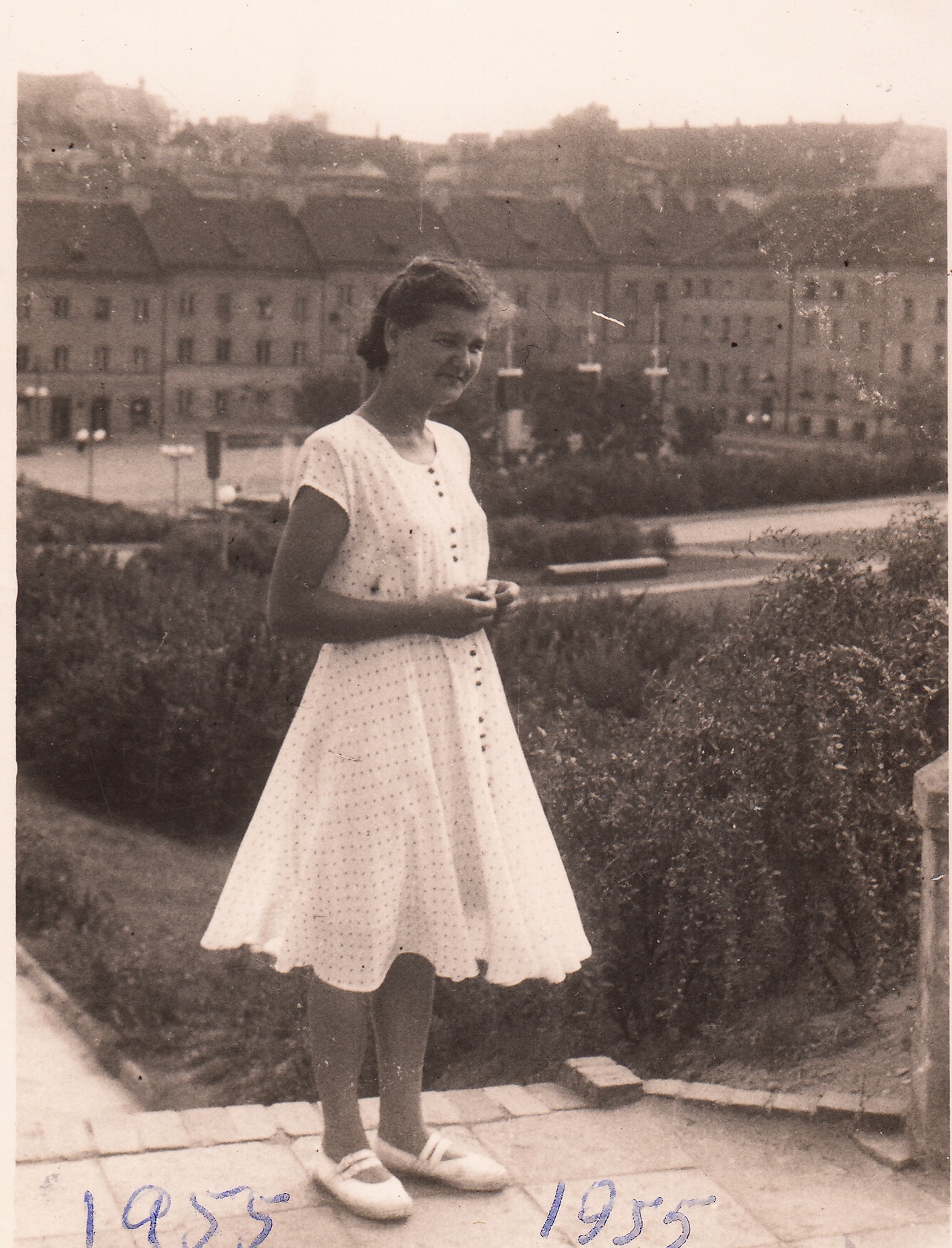 Młoda kobieta w białej sukience stoi na zewnątrz na ścieżce z widokiem na miasto w tle. Na zdjęciu widnieje rok „1955”.