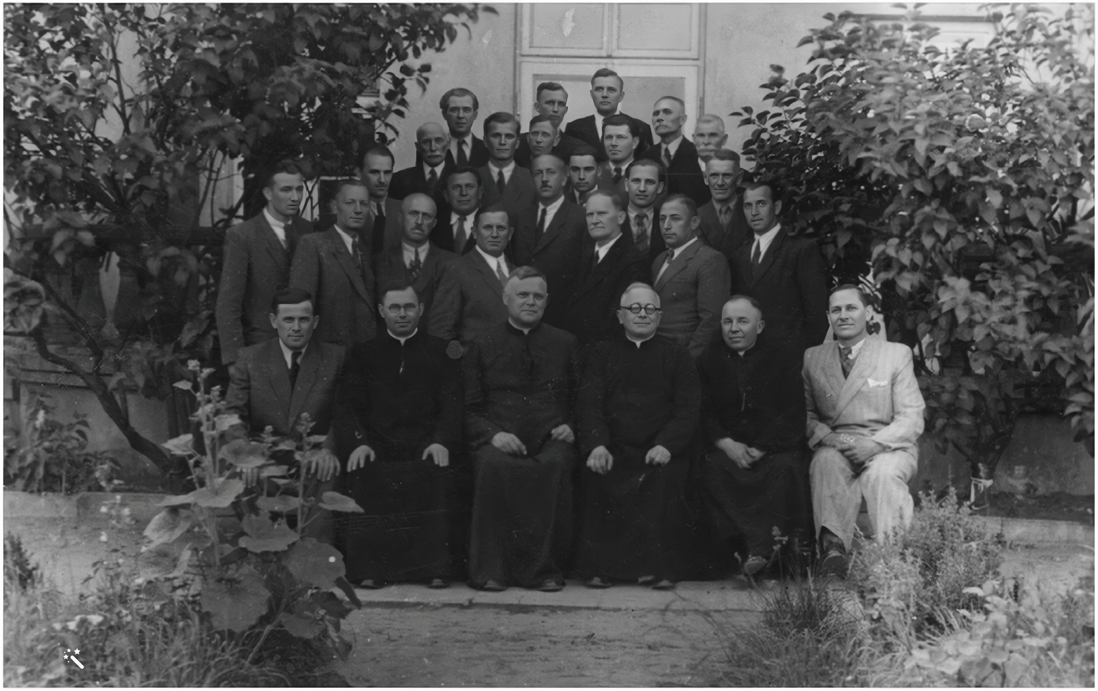 Grupa mężczyzn, niektórzy w strojach religijnych, pozuje do czarno-białego zdjęcia na zewnątrz, przed budynkiem porośniętym roślinnością.