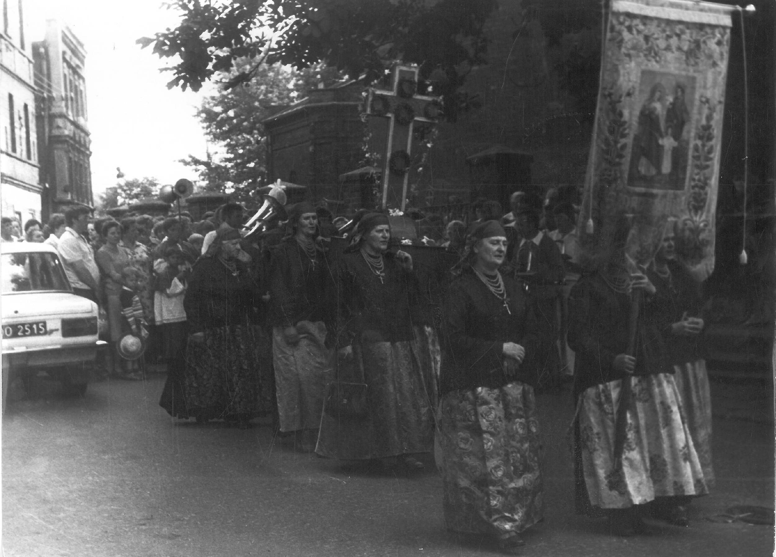 Grupa kobiet w tradycyjnych strojach bierze udział w procesji na świeżym powietrzu, trzymając transparent na ulicy pełnej gapiów i zaparkowanych samochodów.