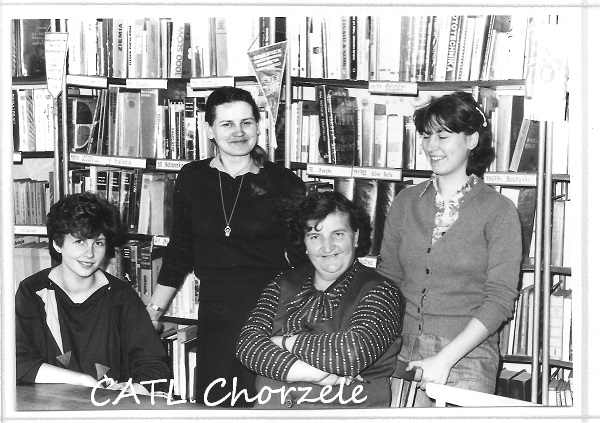 Cztery osoby pozują w bibliotece przed półkami pełnymi książek. Trzy stoją, a jedna siedzi ze skrzyżowanymi ramionami. Na dole ręcznie napisany tekst brzmi „CAFiL Chorzele”.