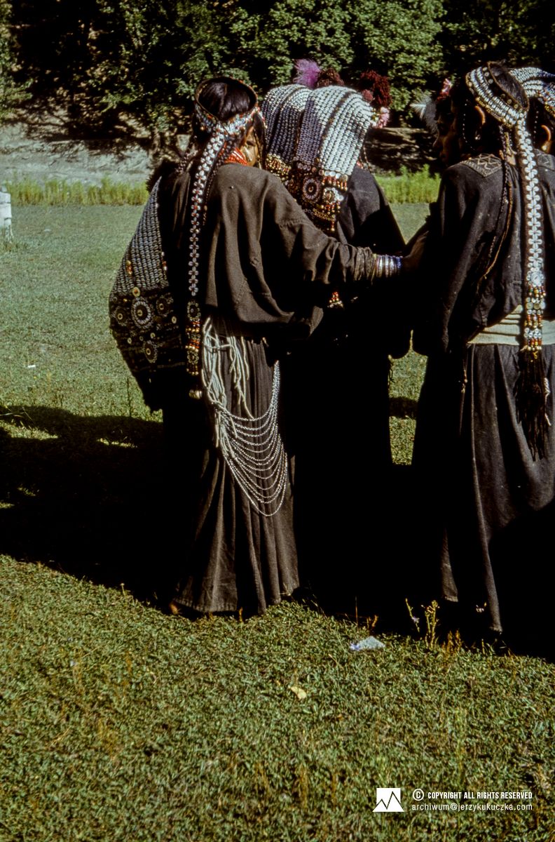 Grupa ludzi ubranych w tradycyjne stroje i nakrycia głowy stoi na trawie, w tle widać drzewa.