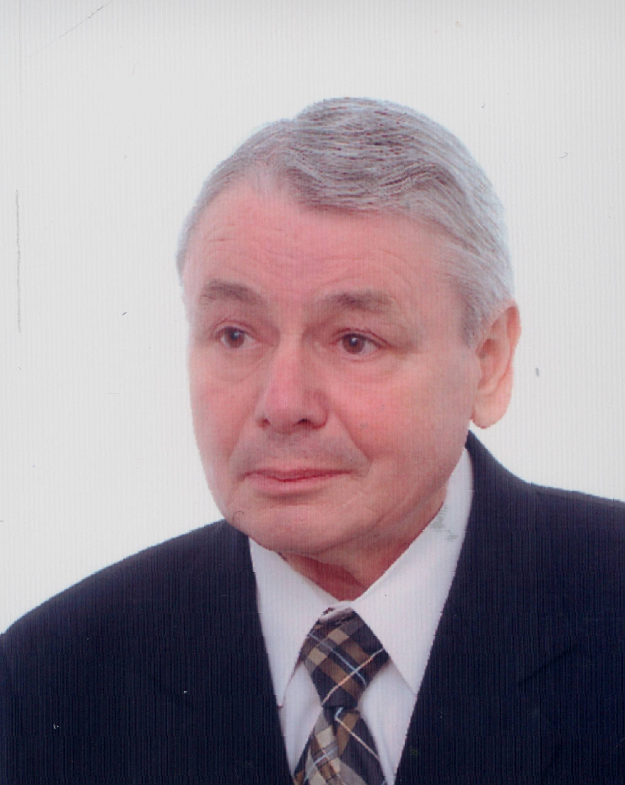 Mężczyzna o siwych włosach, ubrany w garnitur i krawat, patrzy lekko w bok na jednolitym tle.