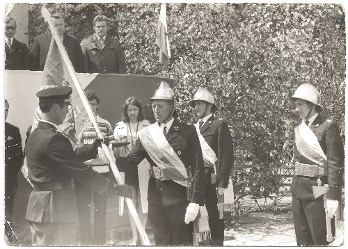 Zdjęcie archiwalne umundurowanych mężczyzn na ceremonii; jeden mężczyzna trzyma flagę, podczas gdy inni stoją na baczność w hełmach i szarfach. W tle widać drzewa.