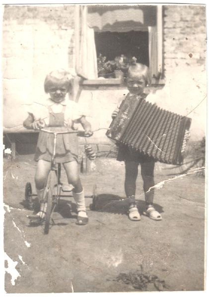 Dwoje dzieci stoi na zewnątrz; jedno na trójkołowcu, drugie trzyma akordeon. Pies wygląda z okna w tle.