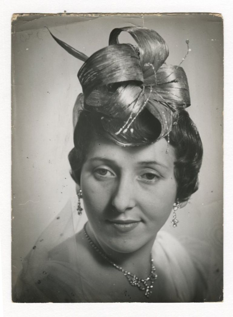 Kobieta w wymyślnym kapeluszu, ozdobiona biżuterią i subtelnym uśmiechem pozuje do portretu na czarno-białej fotografii w stylu vintage.