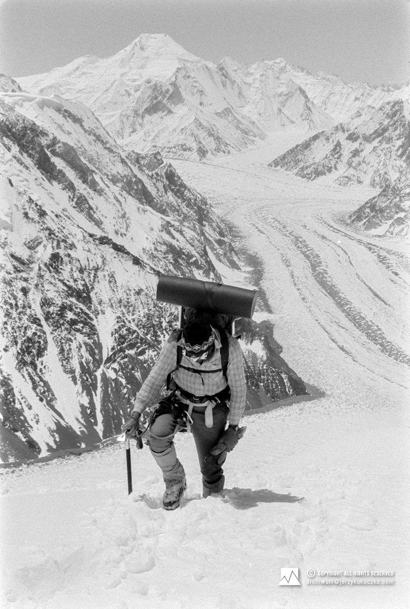 Alpinista wspina się na zaśnieżone zbocze z lodowcem i pasmem górskim w tle, niosąc sprzęt i wspierając się czekanem.