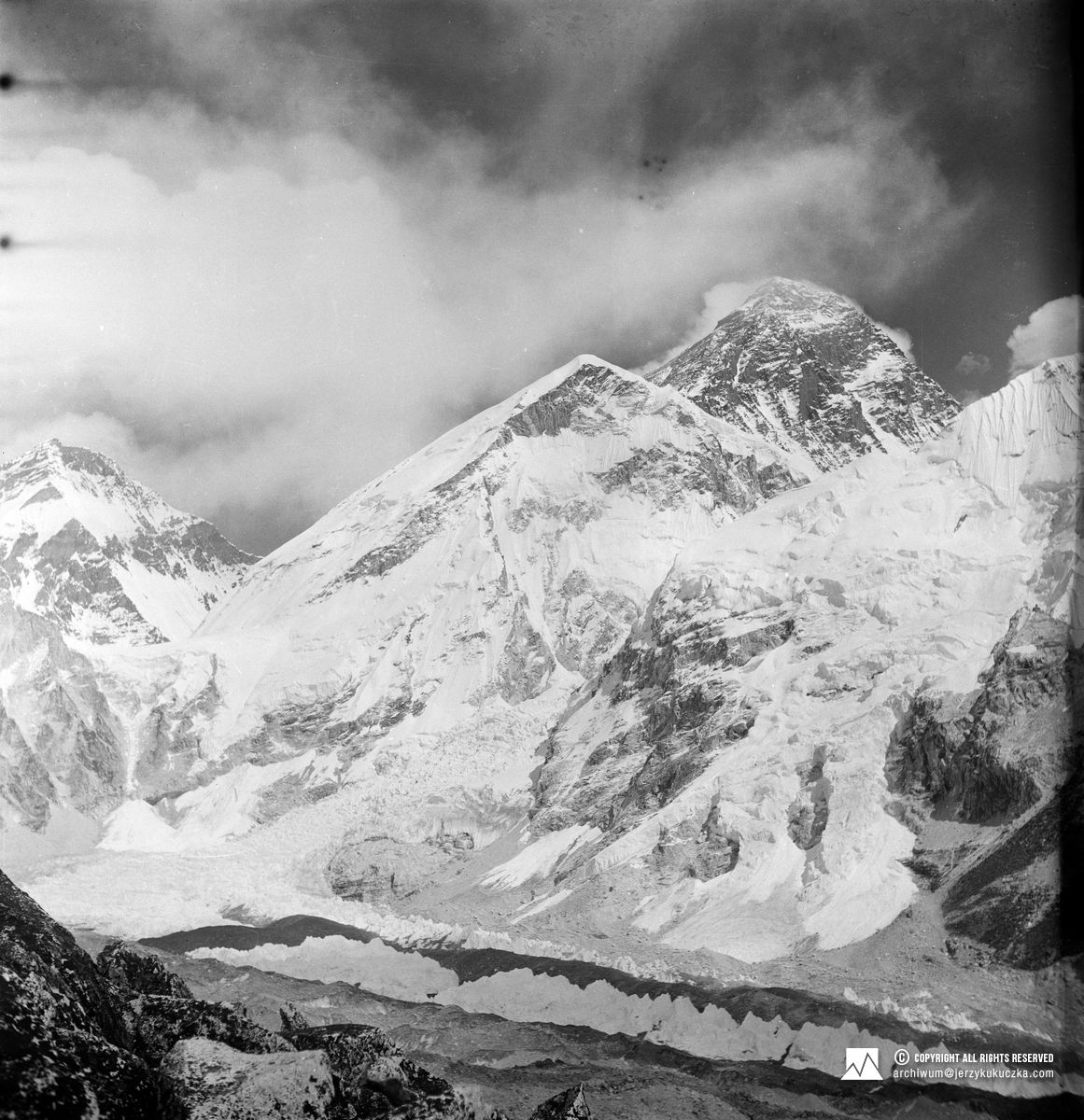 Czarno-białe zdjęcie pokrytego śniegiem Mount Everestu, z chmurami na niebie i lodowcami u jego podnóża.