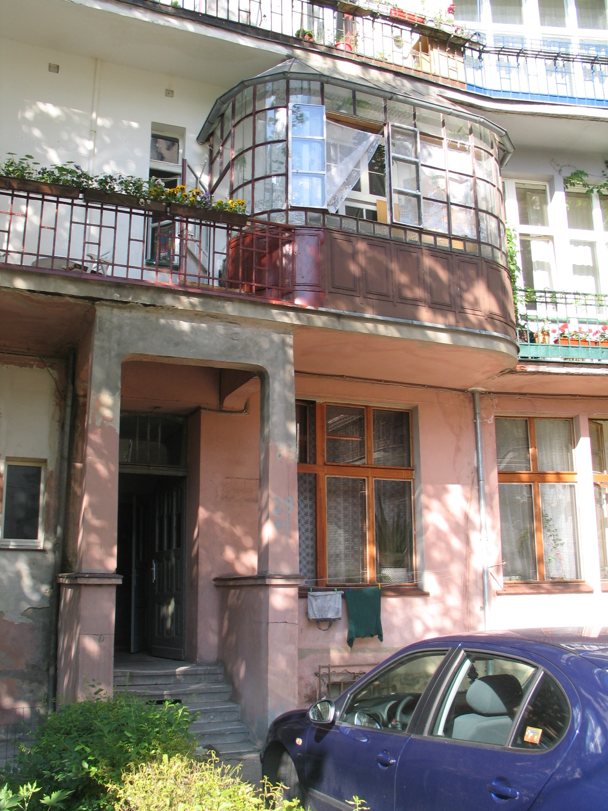 Niebieski samochód zaparkowany jest przed starym budynkiem z przeszklonym balkonem i czerwonymi poręczami. Na balkonie są rośliny, a przy oknie wisi zielona tkanina.