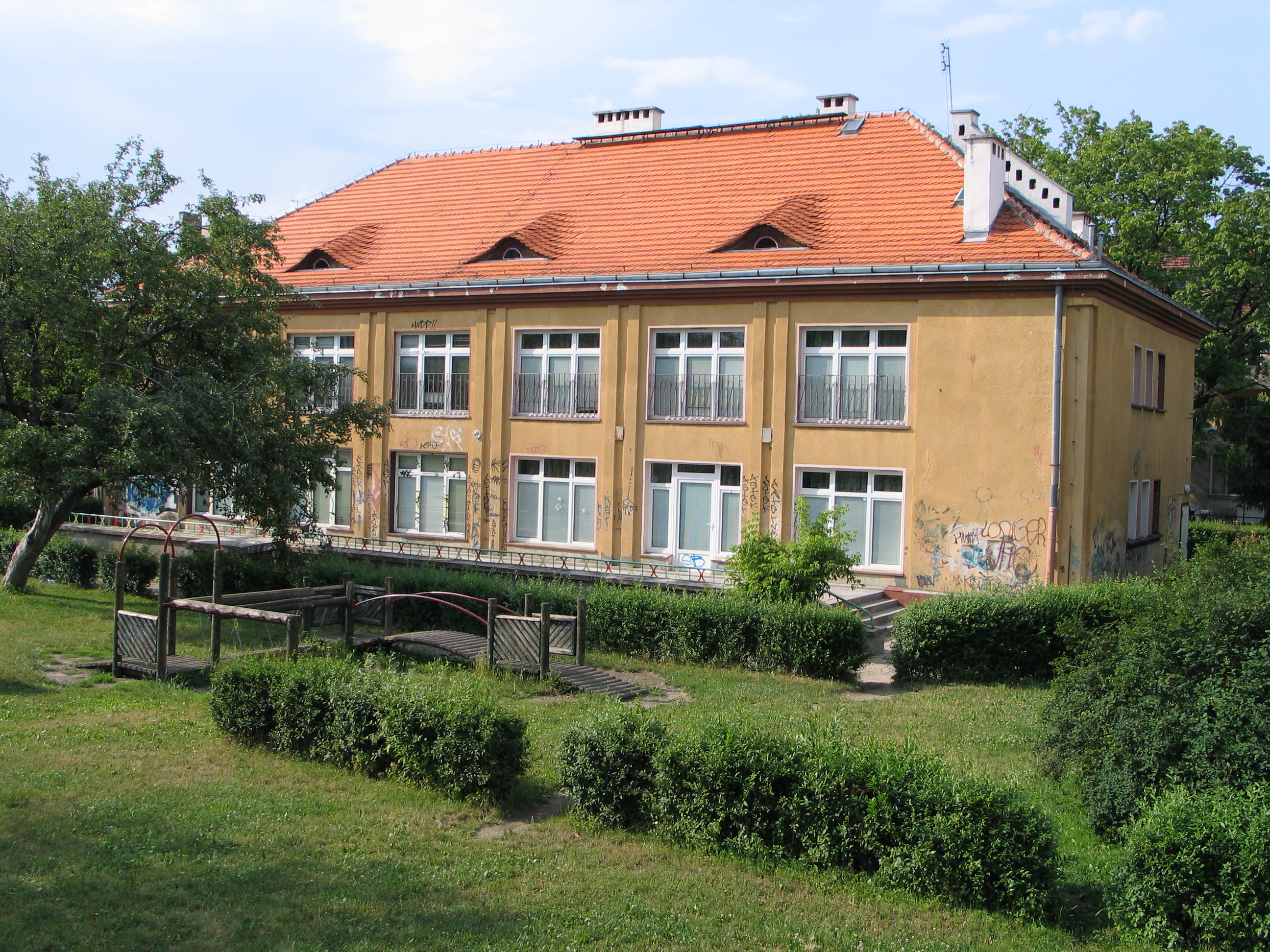 Dwupiętrowy budynek z dachem krytym czerwoną dachówką i licznymi oknami, otoczony zielenią. Na pierwszym planie znajduje się mały plac zabaw.