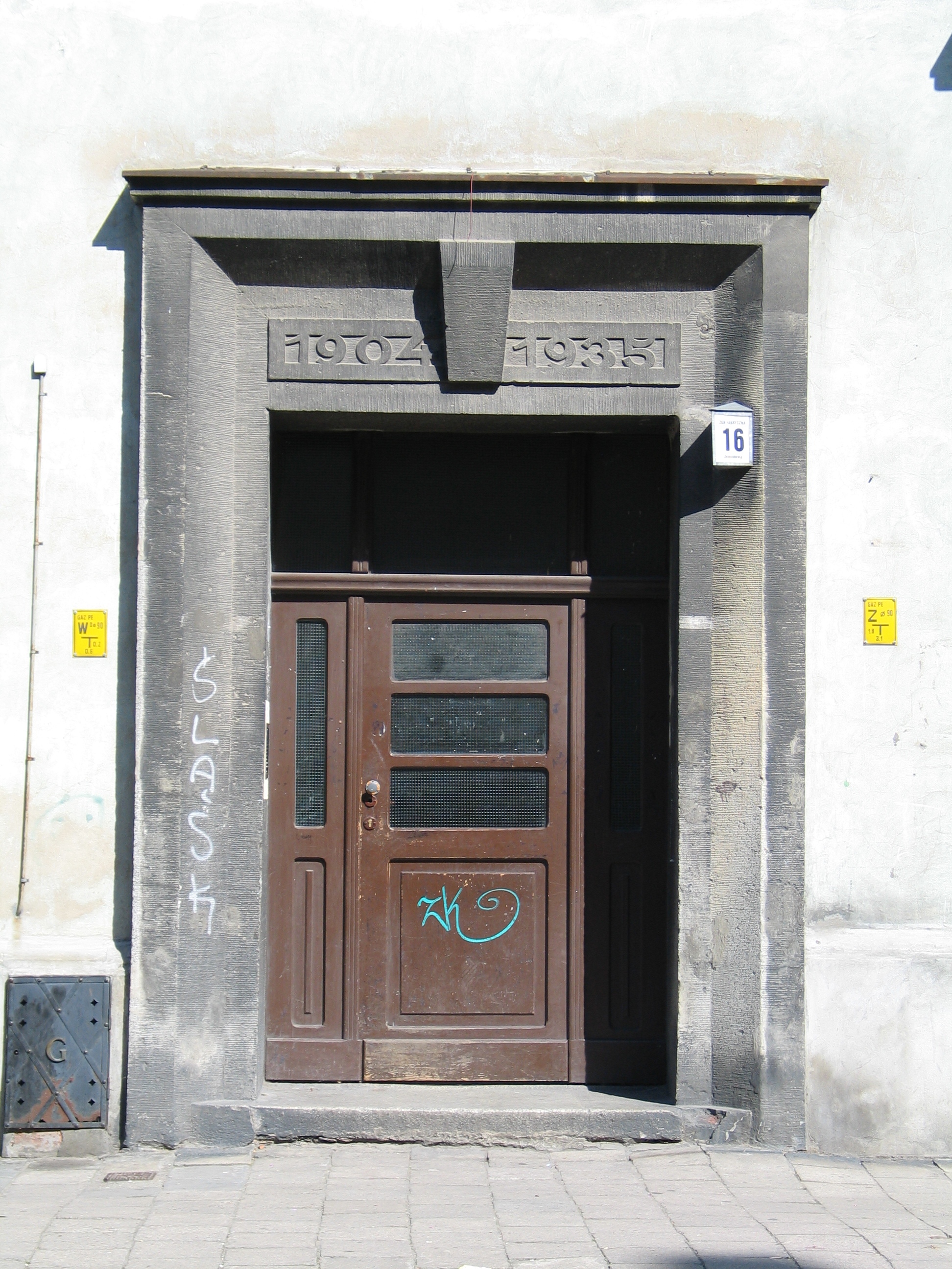 Brązowe drewniane drzwi z panelami szklanymi osadzone są w kamiennej ramie. Rama ma numery u góry i graffiti po bokach. Budynek ma numer 16.