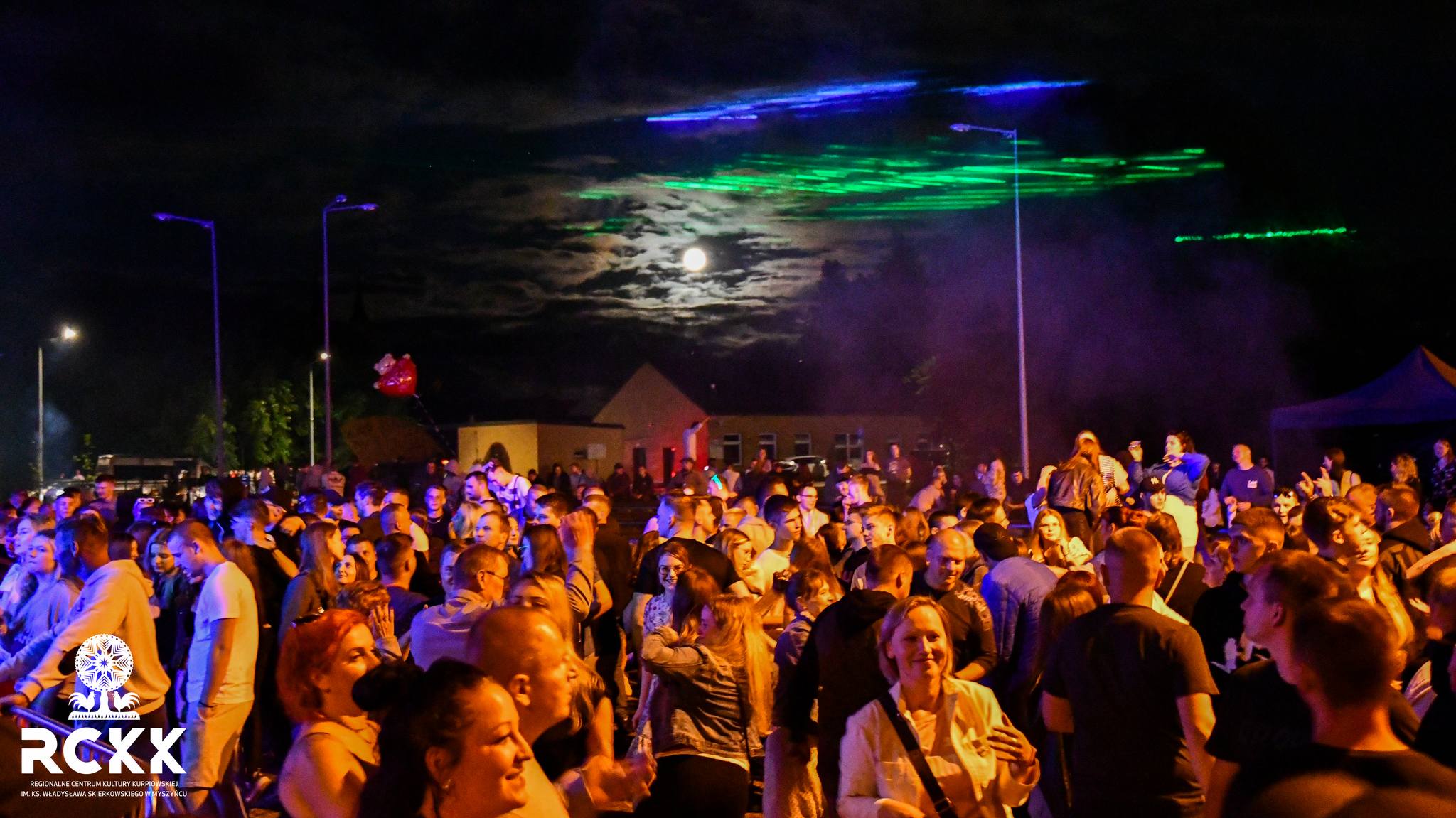 Duży tłum bawiący się na świeżym powietrzu podczas nocnego wydarzenia przy pełni księżyca i kolorowych światłach na niebie.