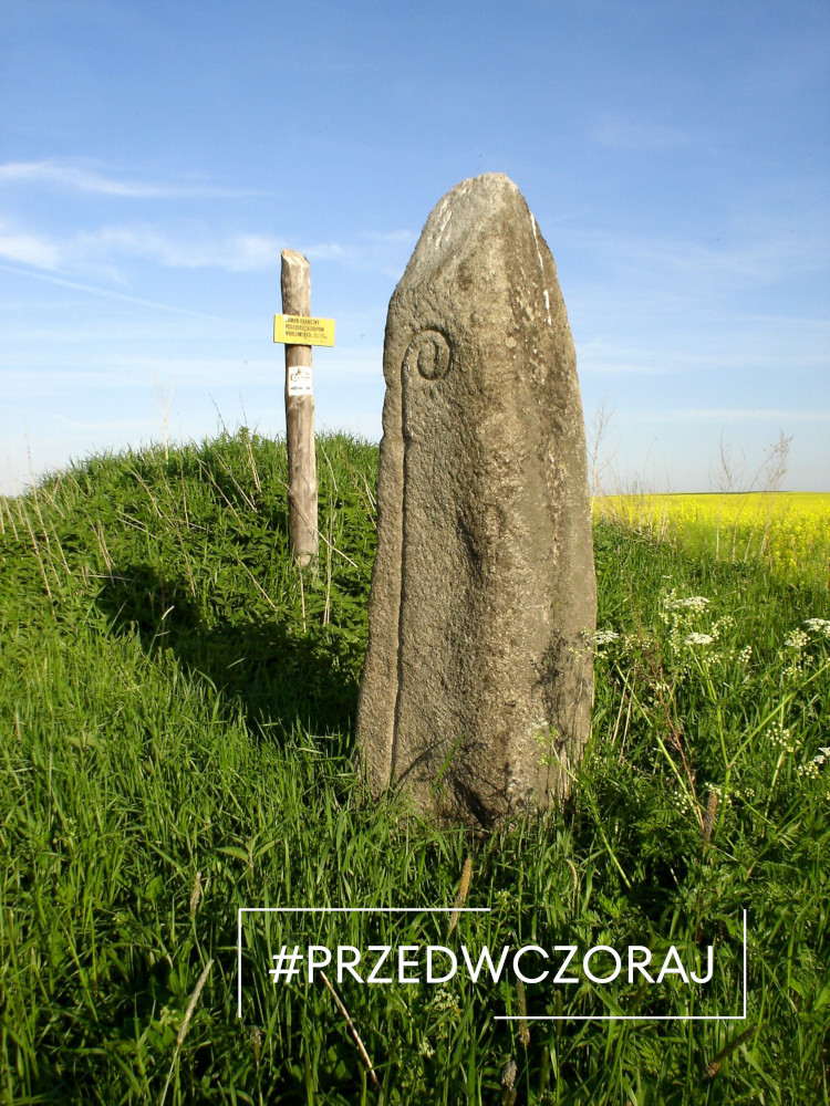 Kamienny pomnik na trawiastym polu z drewnianym słupkiem i żółtym znakiem w tle. Niebieskie niebo nad głową. Nakładka tekstowa brzmi „#PRZEDWCZORAJ”.