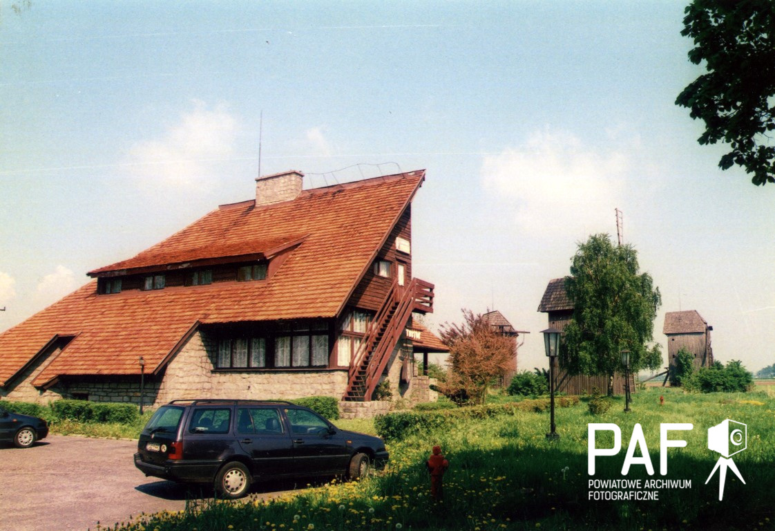 Rustykalny budynek ze stromym, czerwonym dachem stoi w pobliżu zaparkowanego samochodu. Niebo jest częściowo zachmurzone, a obszar otacza pole z dzikimi kwiatami.