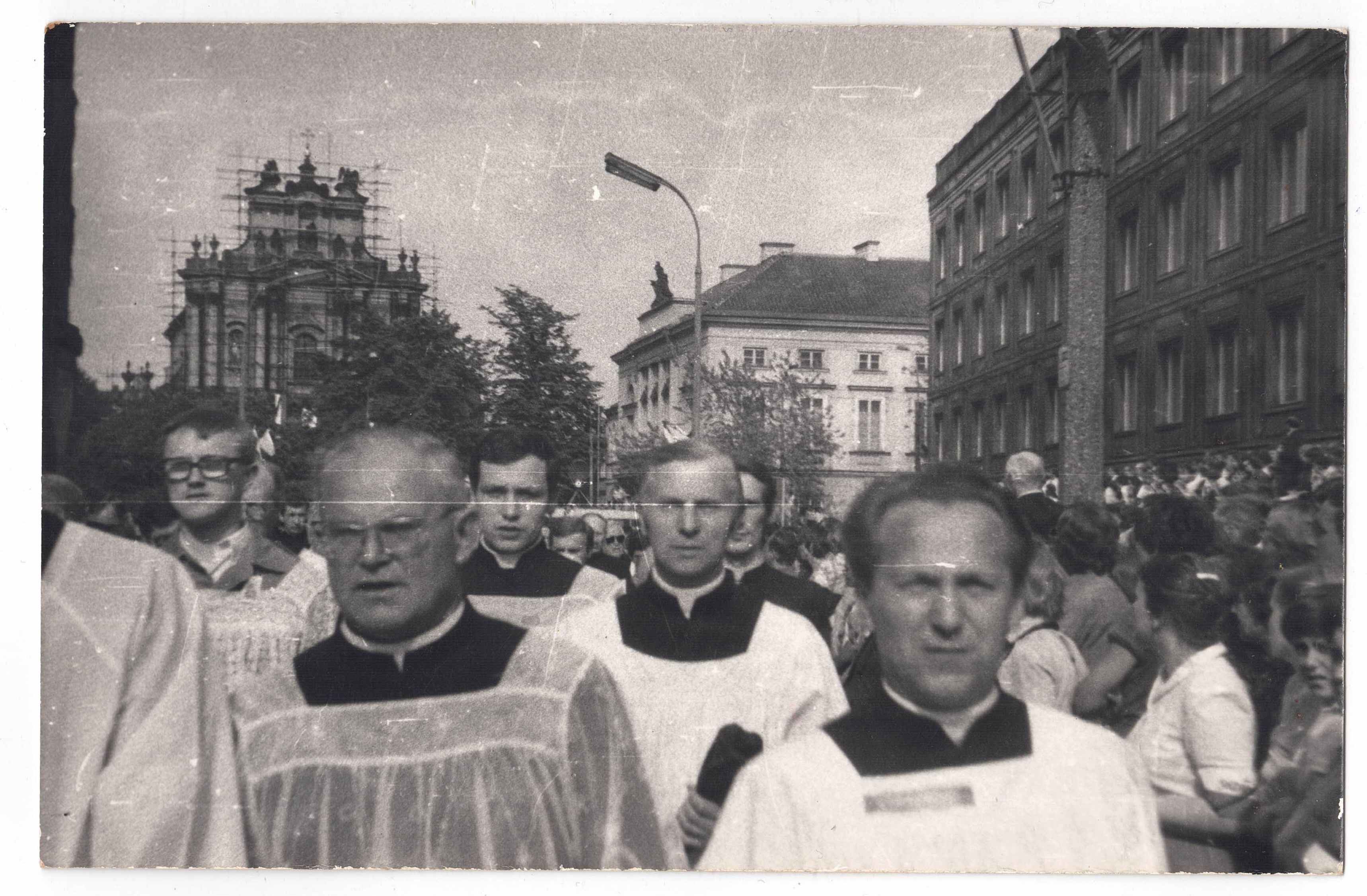 Czarno-białe zdjęcie procesji z duchownymi w tradycyjnych szatach, idących ulicą miasta, otoczonych tłumem. W tle widoczny jest budynek z rusztowaniami.