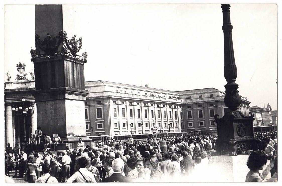 Zatłoczony plac z obeliskiem i ozdobną latarnią na pierwszym planie. Duże budynki i zgromadzenie ludzi wypełniają tło. Obraz jest czarno-biały.