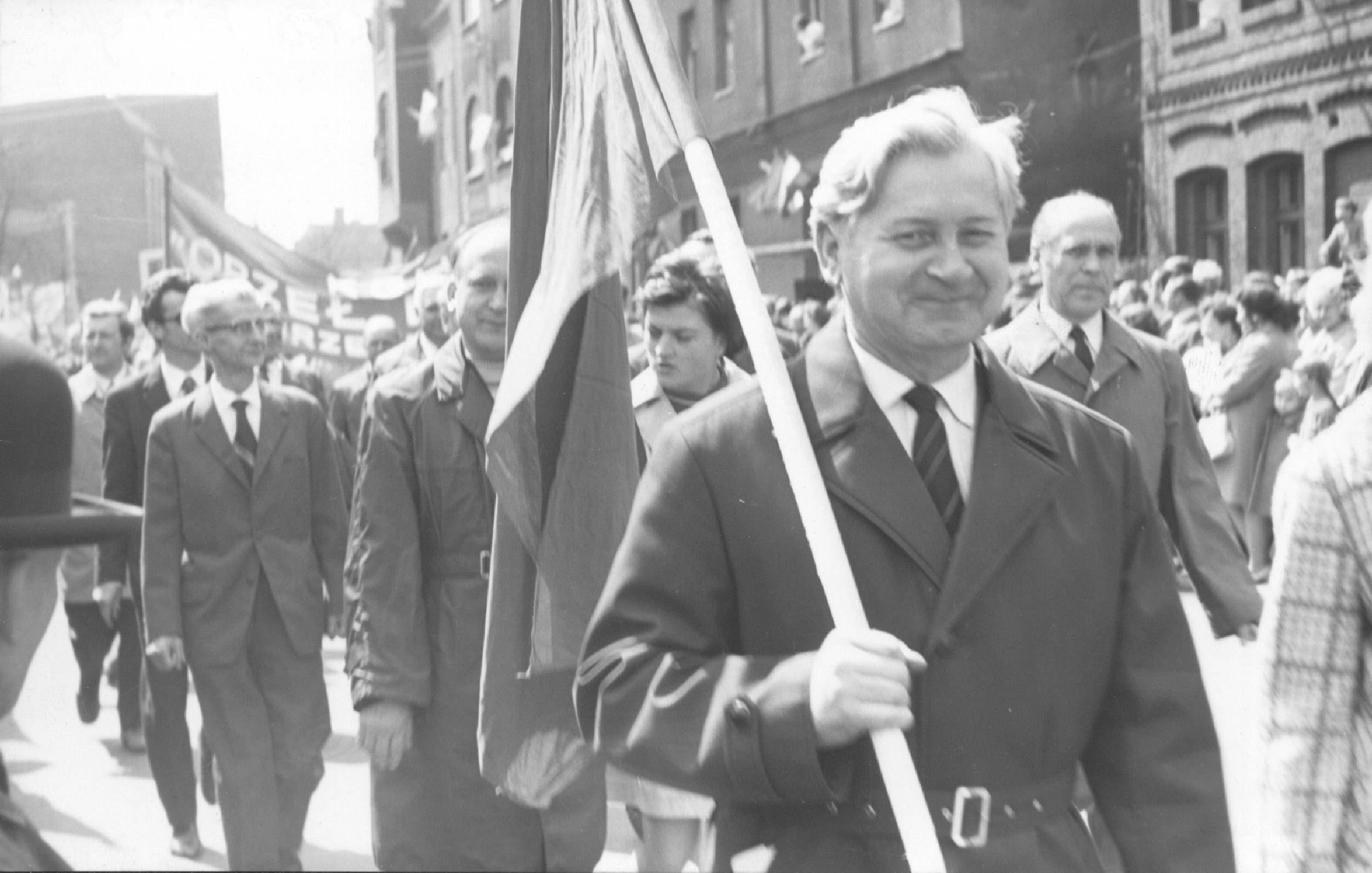 Mężczyzna w garniturze trzyma flagę i prowadzi grupę ludzi maszerujących ulicą. Budynki w tle i obecni są gapie.