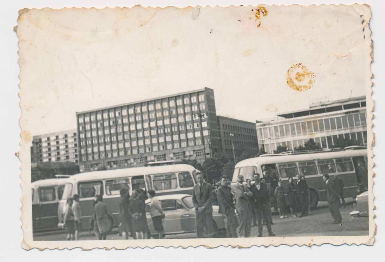 Czarno-białe zdjęcie przedstawiające ludzi stojących obok autobusów na parkingu, w tle widoczne duże budynki biurowe.