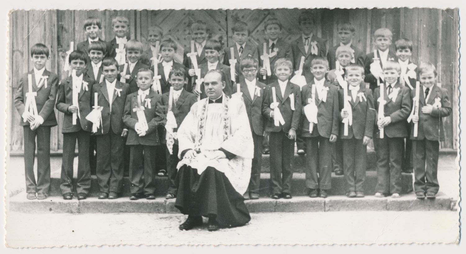 Grupa chłopców w garniturach trzymających świece pozuje z siedzącym księdzem w ceremonialnych szatach przed budynkiem, prawdopodobnie po ceremonii religijnej.