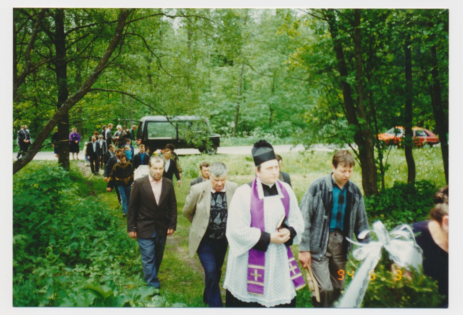 Grupa ludzi, w tym ksiądz w stroju zakonnym, idzie w kondukcie pogrzebowym przez zalesiony teren.