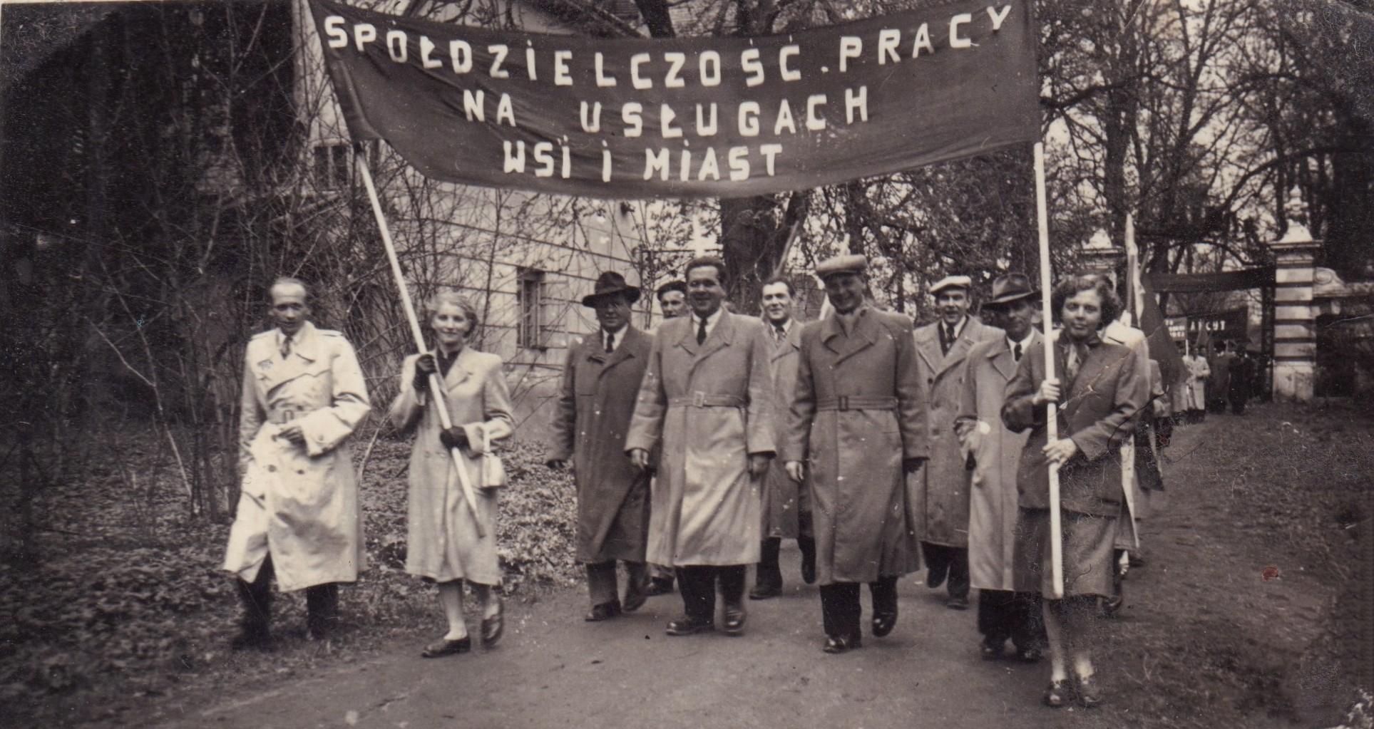 Grupa ludzi w płaszczach kroczy na zewnątrz, trzymając transparent z napisem w języku polskim „Spółdzielczość pracy na usługach wsi i miast”, co oznacza „Spółdzielnie pracy na rzecz wsi i miast”.