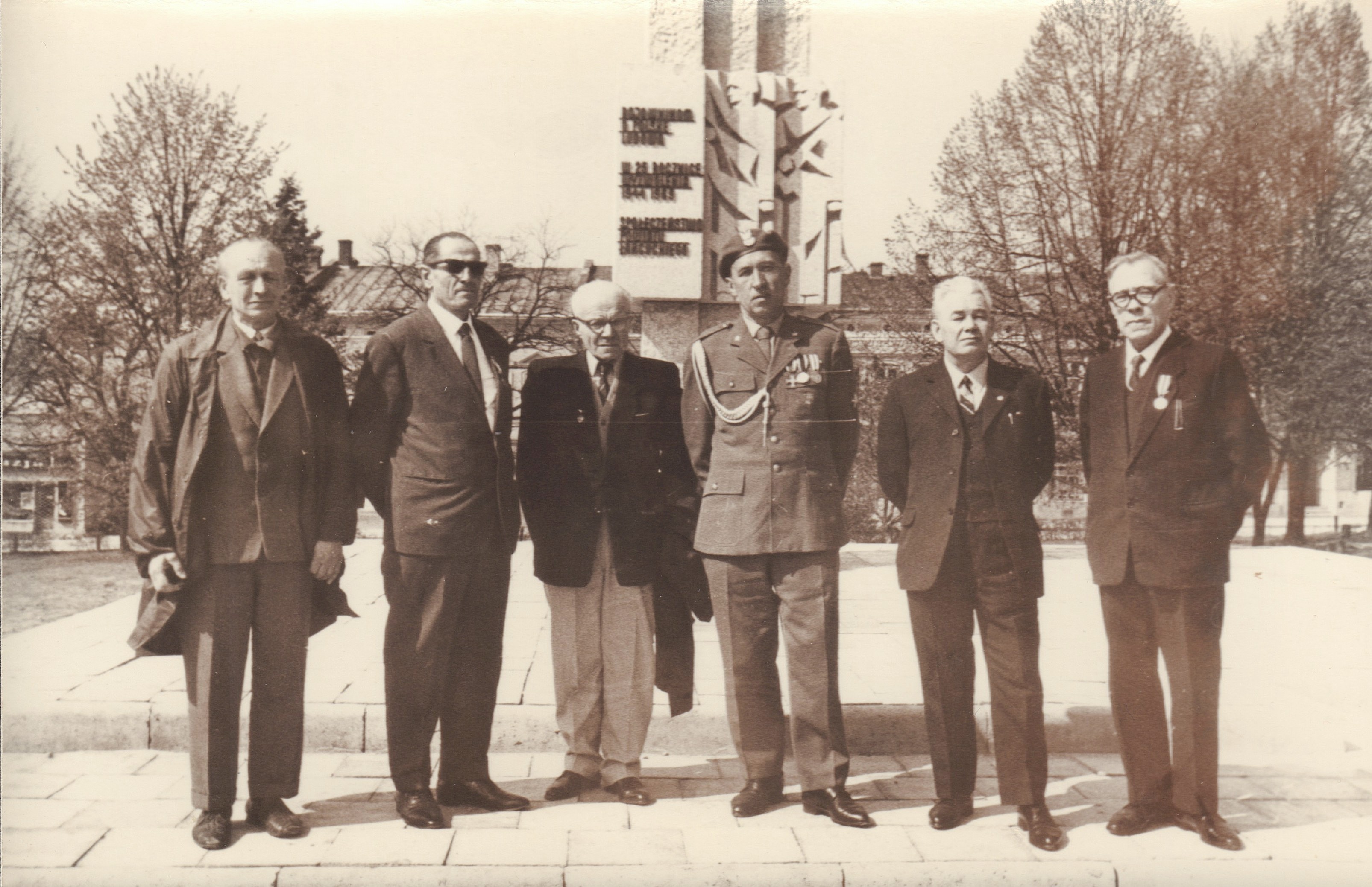 Sześciu mężczyzn, niektórzy w garniturach, a jeden w mundurze wojskowym z medalami, stoi w rzędzie na zewnątrz przed pomnikiem i drzewami.