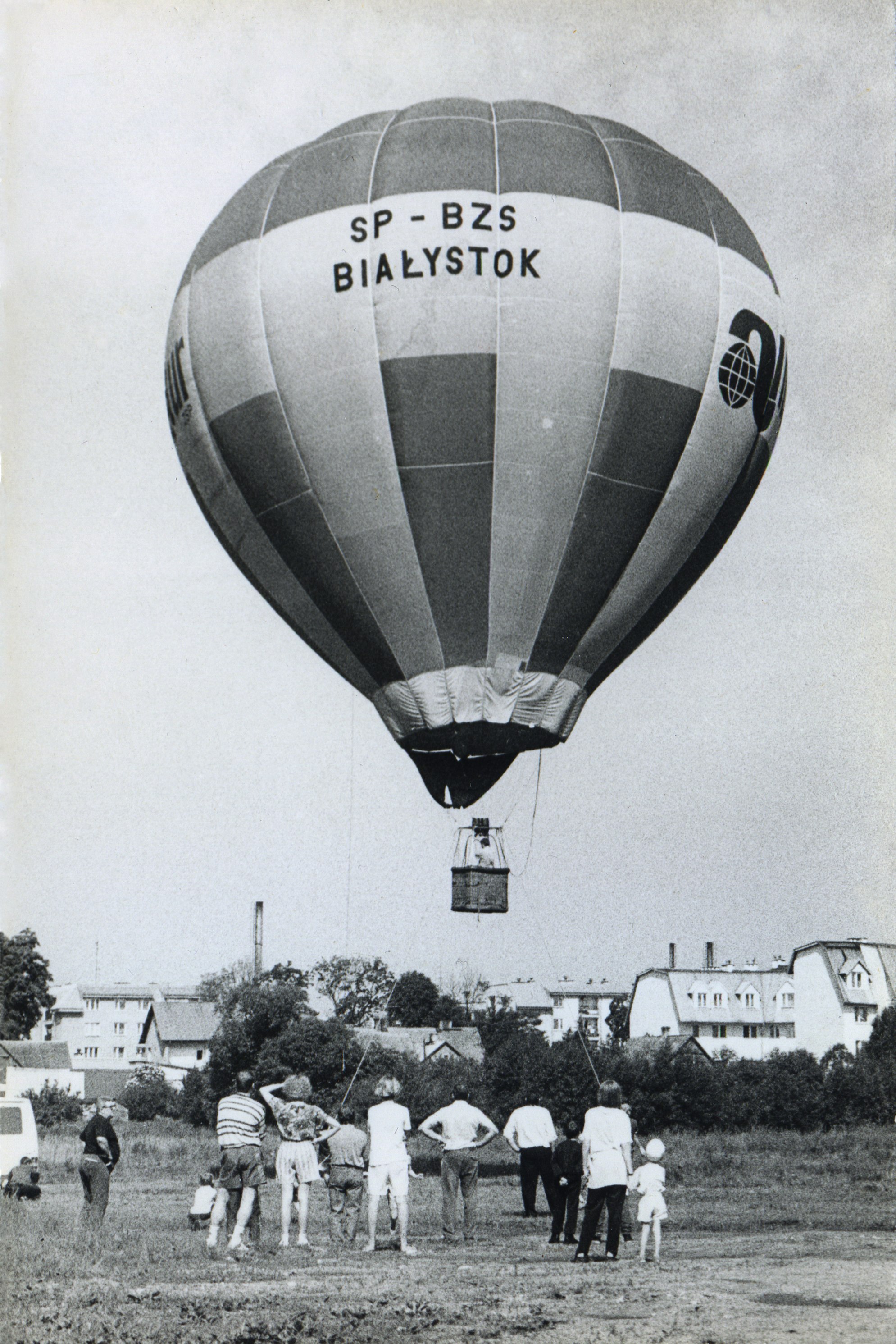 Balon na ogrzane powietrze oznaczony „SP-BZS Białystok” przygotowuje się do startu, podczas gdy grupa ludzi stoi na ziemi i obserwuje. W tle widoczne są budynki mieszkalne.