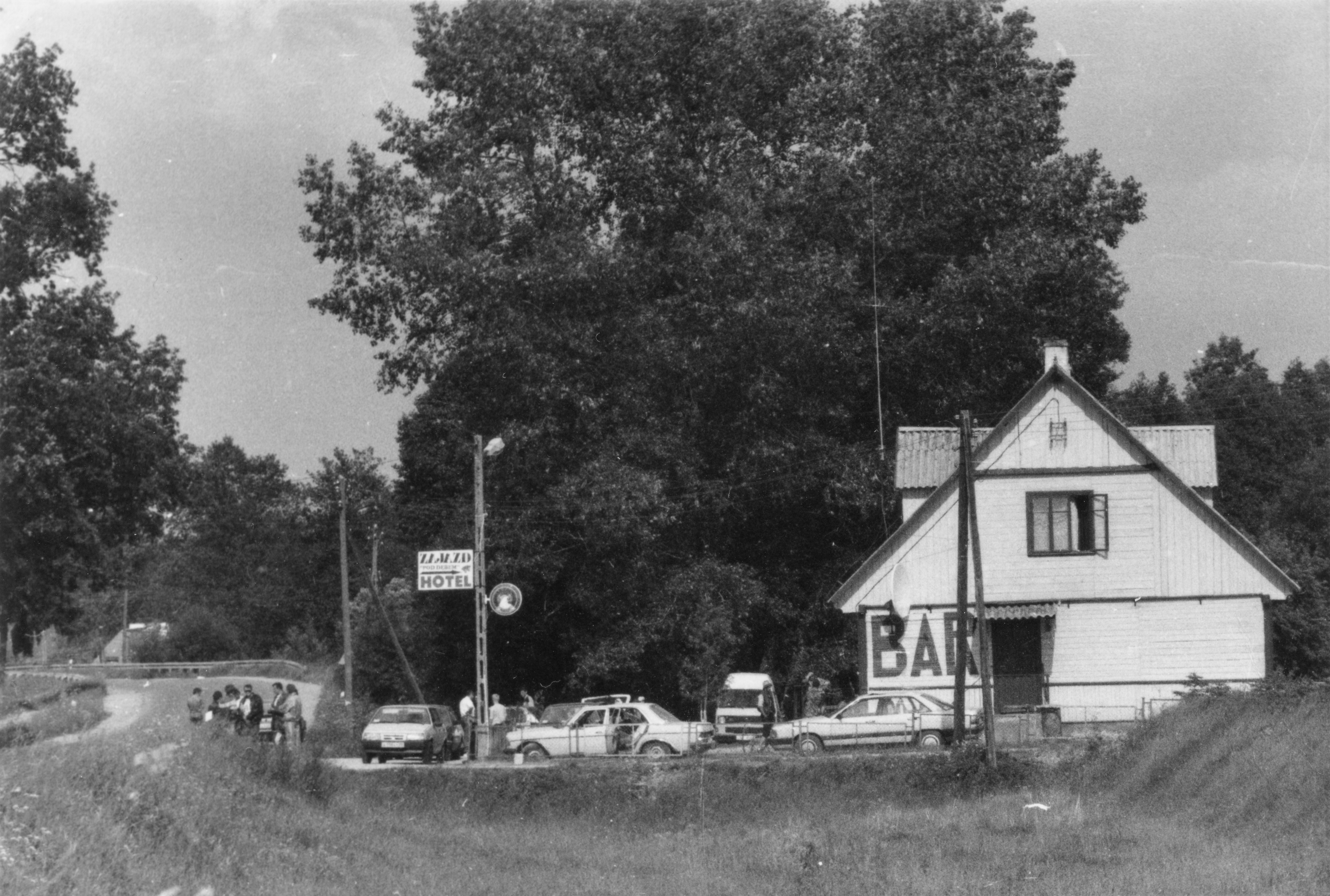 Czarno-białe zdjęcie przydrożnego hotelu ze znakiem „BAR”, kilkoma zaparkowanymi samochodami i ludźmi zgromadzonymi na zewnątrz, pod drzewami.