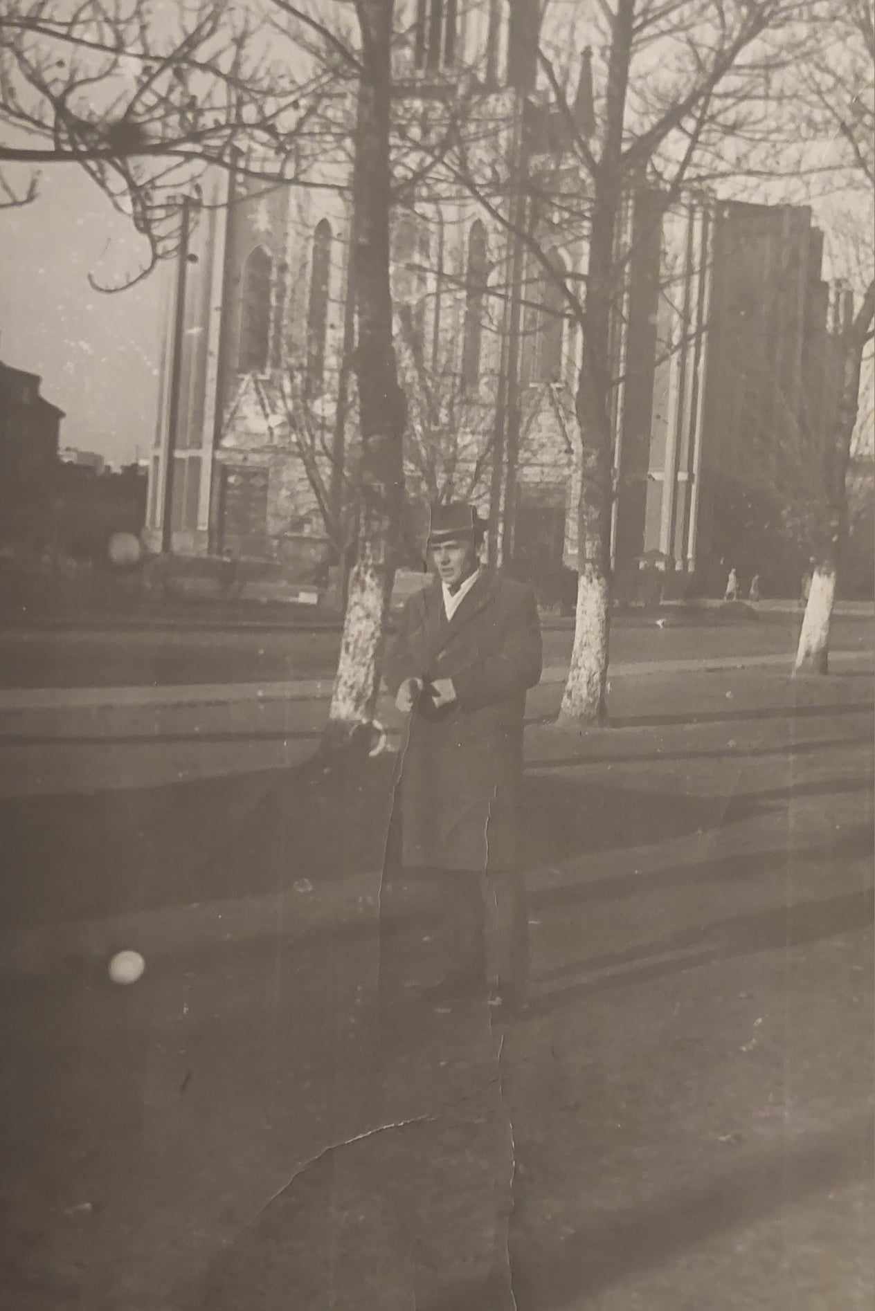 Na czarno-białej fotografii widać mężczyznę w długim płaszczu i kapeluszu stojącego na ulicy przed kościołem z wysokimi oknami i bezlistnymi drzewami.