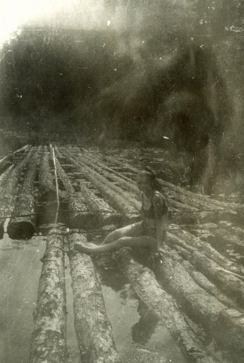 Na czarno-białej fotografii widać osobę siedzącą na grupie dryfujących na wodzie kłód drewna, otoczoną naturalnym otoczeniem.