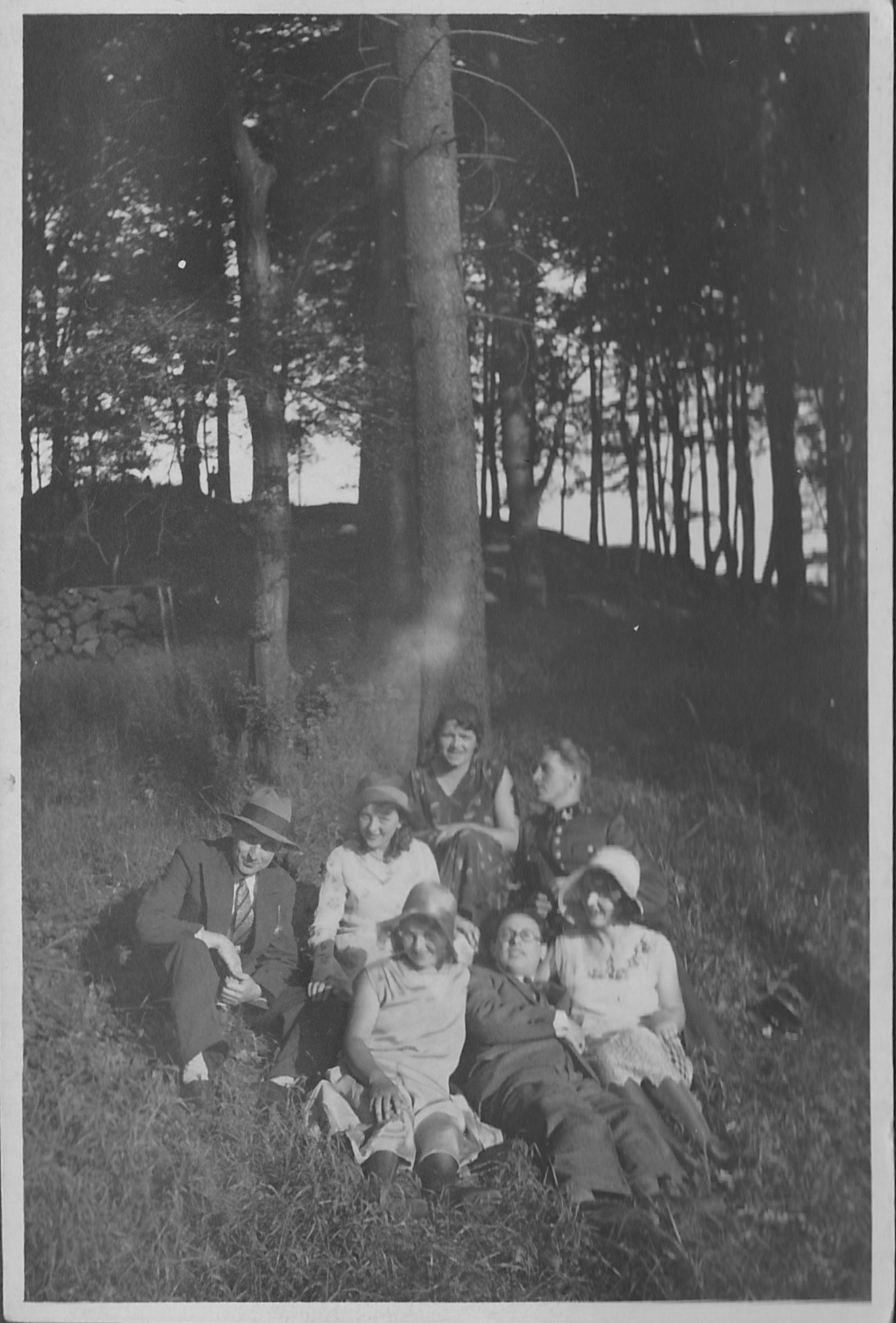 Siedem osób, ubranych w odzież z początku XX wieku, siedzi i odpoczywa na trawie w zalesionym obszarze, z drzewami i światłem słonecznym w tle.