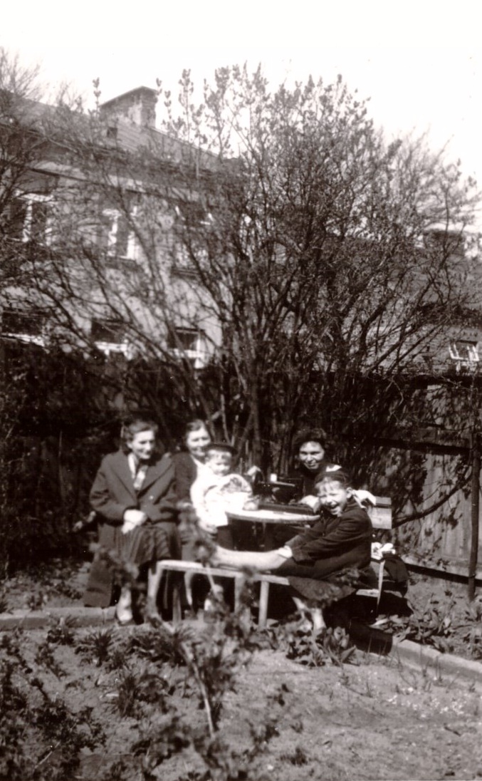 Czworo dorosłych i dziecko zebranych wokół stołu w ogrodzie, z drzewami i budynkiem w tle.