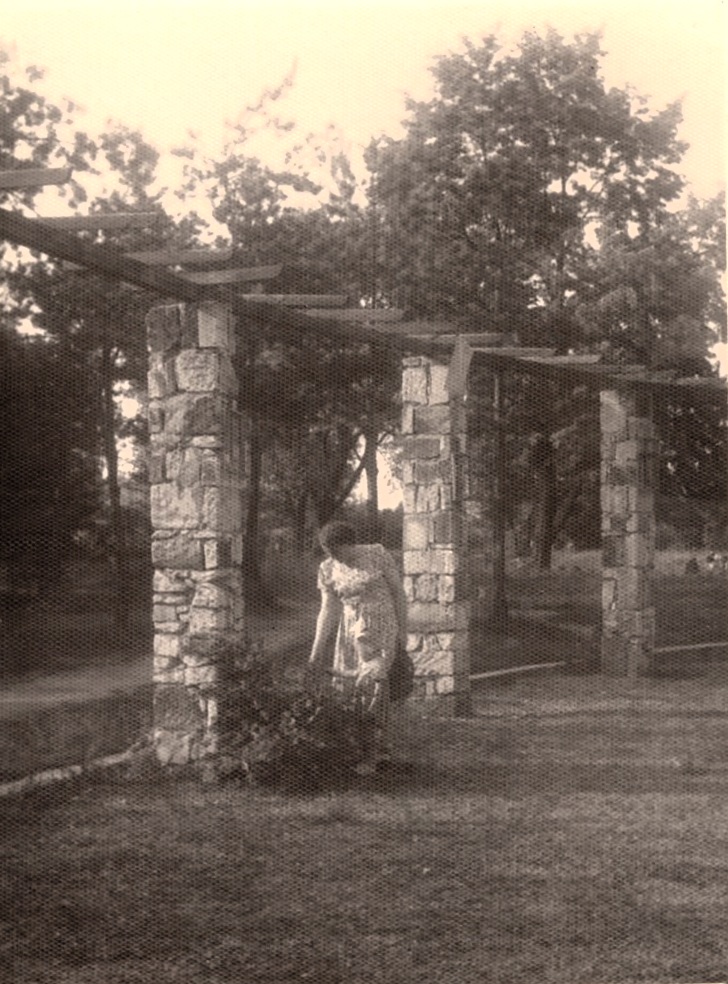 Osoba w sukience stoi obok kamiennej kolumny na trawiastym terenie z drzewami w tle.