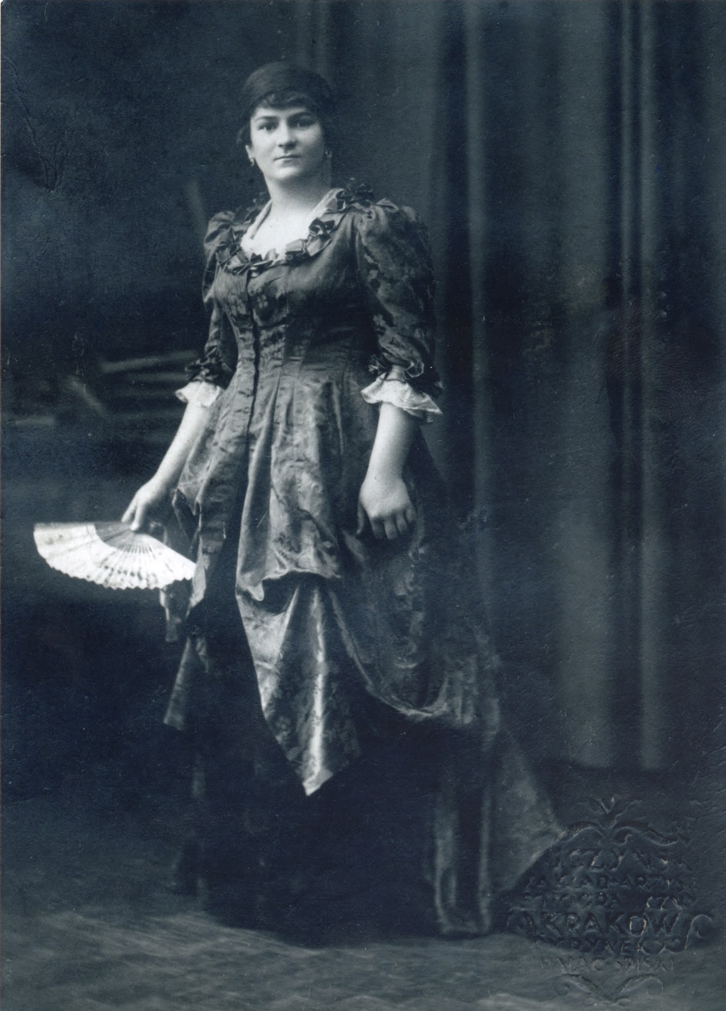 Kobieta w ozdobnej, długiej sukni trzyma wachlarz i stoi przed ciemną zasłoną na tym czarno-białym zdjęciu w stylu vintage.