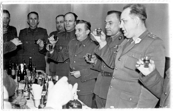 Grupa oficerów w mundurach wznosi toast przy stole pełnym butelek i kubków.